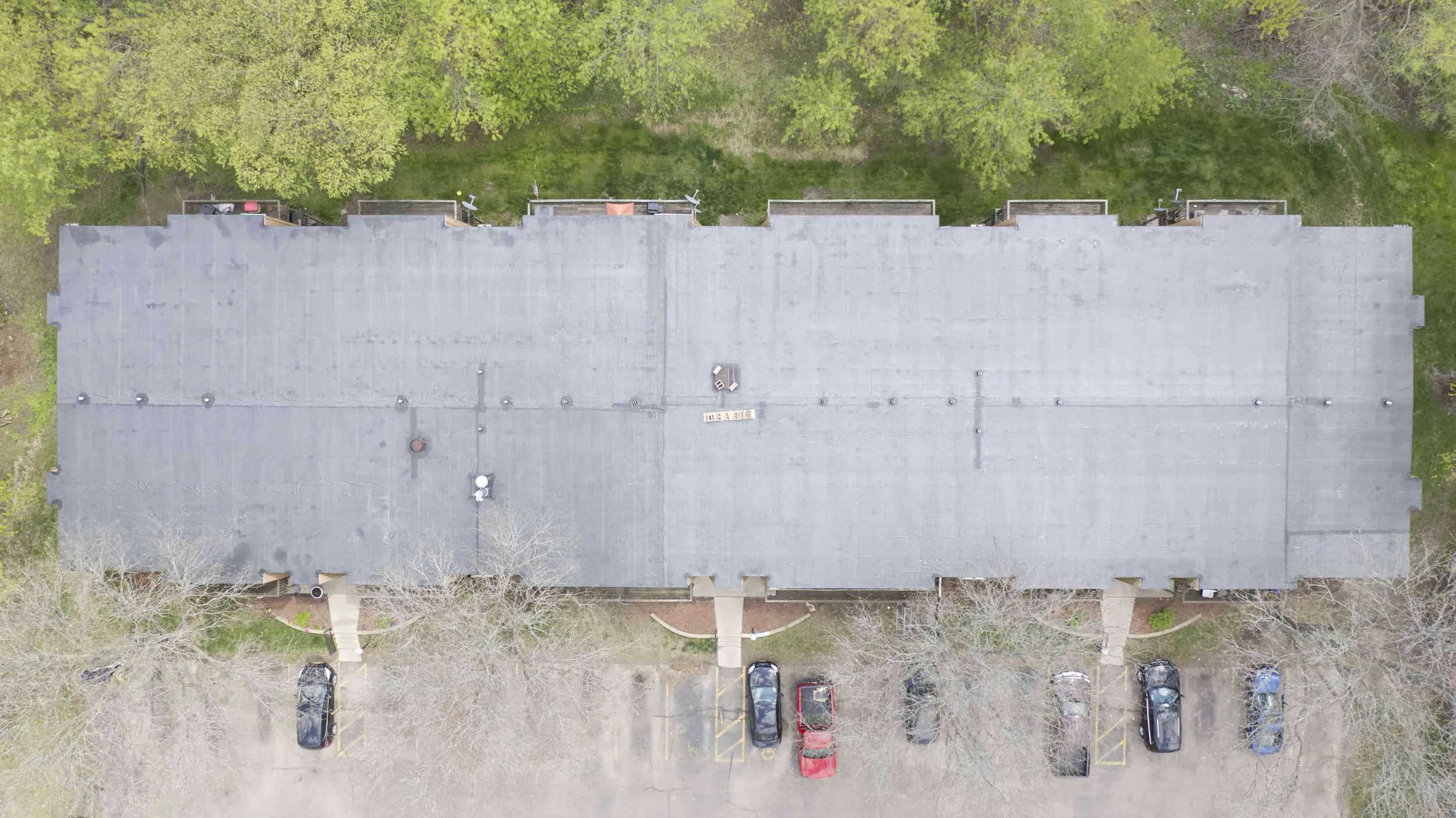 Aerial view of a rectangular apartment building with a gray flat roof, surrounded by green trees and a parking lot with several cars.
